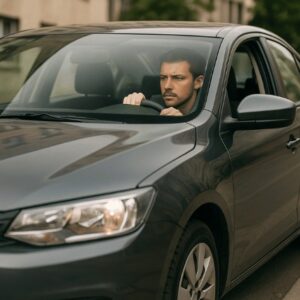 Pessoa dirigindo um carro cinza sem placa em uma rua urbana, vista lateral e em plano médio, com expressão concentrada e iluminação suave natural