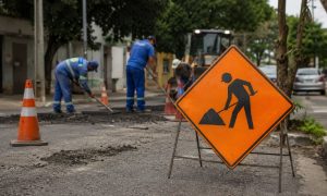 rua em manutenção, com placa de obras em destaque no primeiro plano e trabalhadores consertando o asfalto ao fundo, em um cenário urbano informativo e visualmente envolvente