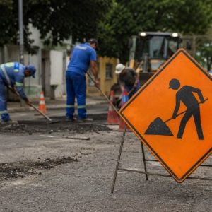 rua em manutenção, com placa de obras em destaque no primeiro plano e trabalhadores consertando o asfalto ao fundo, em um cenário urbano informativo e visualmente envolvente